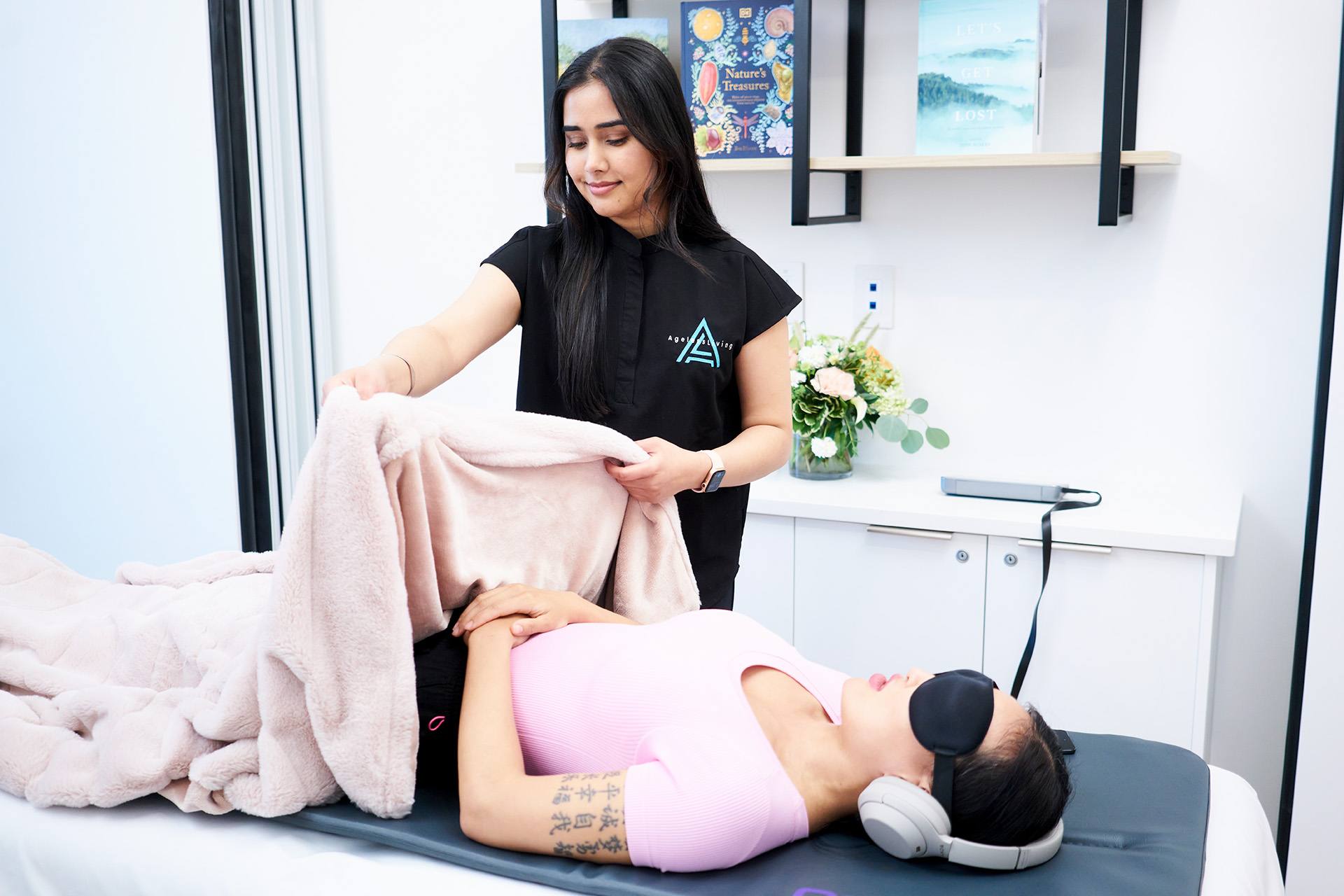 An Ageless Living practitioner settling a resting client under a blanket, in an eye mask and headphones